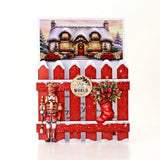A square Christmas greeting card featuring a snowy winter house illustration above a red picket fence with a nutcracker, a red stocking, and a round badge that reads 'Joy of the WORLD'.