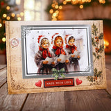 A decorative handmade Christmas greeting card displayed on a wooden surface with a blurred festive background. The card features a rustic beige border with ornate scrollwork, a central photo of three young girls in winter clothing holding sheet music, red hearts, a green bow, and a red banner that reads 'MADE WITH LOVE'.