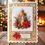 A handmade Christmas greeting card with a scalloped white border, featuring an illustration of a lantern, red poinsettias, pinecones, and vintage postage-style stamps. The card is displayed on a wooden surface with a warm, blurred holiday background.
