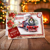 A Christmas-themed greeting card featuring a vintage steam locomotive, red poinsettias, and holiday text, displayed on a wooden surface with a warm, blurred fireplace background.