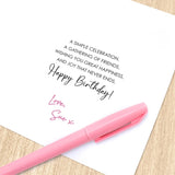 A white birthday card with printed black text and a pink handwritten signature, placed on a light wood surface with a pink pen resting across the lower part of the card.