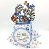 A 3D pop-up dog birthday card with a blue and white striped base decorated with bone and paw print motifs. The card features a central blue scalloped label with a message for a happy birthday, and rising from the base are cartoon gifts wrapped in red, gold, and blue ribbon, topped with treats and banners reading 'DOGGY' and 'WOOF'.