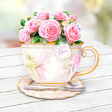 Decorative teacup with pink roses and a 'With Love' tag on a wooden surface.