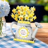 A decorative watering can-shaped card or gift holder filled with yellow daffodils, placed on a blue-and-white checkered tablecloth beside a glass vase of blue flowers. The front has a round label reading “Love & Birthday Wishes.