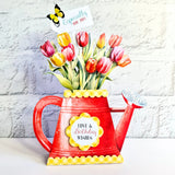 A red watering can shaped vase filled with colorful tulips, featuring a yellow scalloped trim, a flower pot emblem with 'Love & Birthday Wishes', and a butterfly tag that reads 'Especially for you!'.