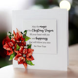 A white greeting card with a printed holiday message, decorated with a raised red poinsettia and green leaves on the left side, photographed on a softly blurred indoor background.