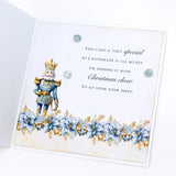 An open white greeting card featuring a decorative nutcracker illustration in blue and gold, blue poinsettia flowers along the bottom, and printed holiday text on the front.