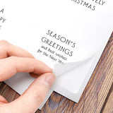 A close-up of a hand peeling back a transparent protective film from a white paper holiday greeting card placed on a wooden surface. The card shows printed text including 'SEASON'S GREETINGS' and partial 'MERRY CHRISTMAS'.