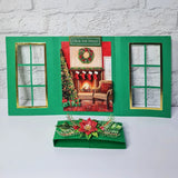 A green Christmas-themed card game set displayed on a white surface against a white brick wall. The set includes an open triptych-style card frame with gold-trimmed window cutouts and a central holiday scene, plus a matching green box with a red poinsettia and green leaf embellishment.