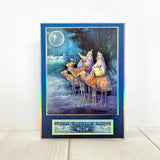 A vertical art print or card featuring the Three Wise Men under a full moon, with a holographic iridescent blue border and a small matching strip at the bottom, displayed on a light wood surface against a white wall.