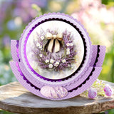 A layered circular card or decorative plaque with purple scalloped borders and a central floral wreath photo, displayed on a rustic wooden surface with blurred pink flowers in the background.