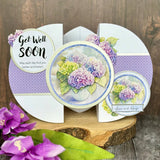 A handmade greeting card displayed on a rustic wood slice, featuring purple and lavender hydrangea illustrations, a purple polka dot pattern, and text reading 'Get Well Soon' and 'Love and Hugs'.
