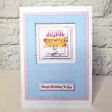 A handmade birthday card featuring a layered blue and pink paper design with a central illustration of a yellow cake and the text 'Happy Birthday To You' at the bottom, displayed against a white brick wall background.