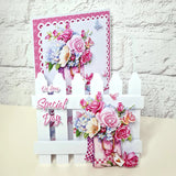 A handmade greeting card displayed on a white picket fence stand against a white brick wall. The card features a pink and white patterned border, a bouquet of pink and blue flowers, butterflies, and the text 'On Your Special Day'.