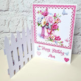 A handmade 3D pop-up birthday card for 'Mum' featuring a pink and white floral bouquet in a checkered vase, a white picket fence on the left, and 'Happy Birthday Mum' written in pink script on a white background.