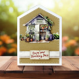 Greeting card with a garden shed and plants on a wooden surface with a blurred floral background.
