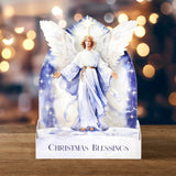 A decorative Christmas card or display stand featuring an angel figure with white wings and a blue robe, standing on a base printed with the words 'CHRISTMAS BLESSINGS.' The background shows warm blurred holiday lights and a wooden surface.