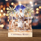 A 3D papercraft Christmas nativity scene display featuring a wise man in white robes standing with three sheep, flanked by palm trees under a starry night sky, set on a wooden base printed with 'CHRISTMAS WISHES'.