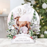 A festive Christmas tabletop decoration featuring a brown deer with antlers walking among snowy trees and holly, with a white banner at the bottom reading 'SENDING SPECIAL CHRISTMAS WISHES'. The decoration is displayed on a light surface with a blurred Christmas tree in the background.