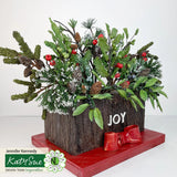 Decorative floral arrangement with greenery, pinecones, and berries on a wooden block with 'JOY' text, on a white background.