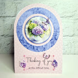 A handmade sympathy card with an arched top and a pink patterned base. The center features a circular layered design with blue paper flowers, a purple hydrangea, green hydrangea, and a butterfly.