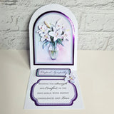 A handmade pop-up condolence card featuring white lilies in a vase, displayed open against a white brick wall background. The card includes a smaller pop-up tab reading 'Roght Sympathy' and a main message of condolence on a white card base with purple borders.