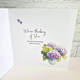 A white greeting card shown open against a white brick wall background. The card features the text 'We're thinking of You and hope you're feeling better very soon.