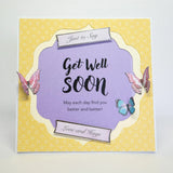 A square greeting card with a yellow patterned border and a central lavender scalloped panel that reads 'Get Well Soon' with smaller text below. Decorative butterfly illustrations appear on the card, along with small ribbon-style labels reading 'Just to Say' and 'Love and Kugs.