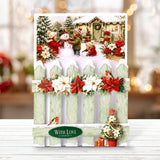 A festive Christmas greeting card displayed on a white wooden surface. The card features a snowy winter scene with a snowman, children, a decorated cabin, poinsettias, and a light green picket fence.