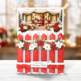 A festive Christmas greeting card featuring a snowy red picket fence with white poinsettias and green garlands, topped with a photo of Santa Claus at a decorated holiday porch. The card is displayed upright on a white wooden surface with a soft, blurred holiday background.